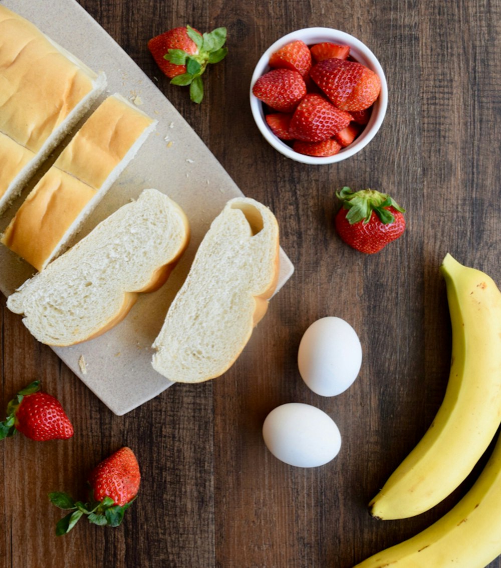 sliced bread on brown wooden chopping board beside yellow banana and red strawberries
