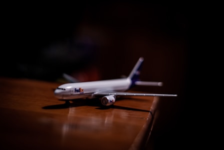A small model airplane with FedEx branding is placed on a wooden surface. The lighting is focused on the plane, with the background in shadow, giving it a dramatic effect.