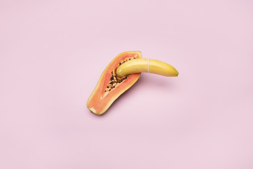 A banana is placed inside a halved papaya with a pink background. The papaya seeds are visible, and the banana is secured with a rubber band.