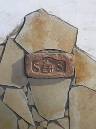 A section of a stone or brick wall featuring a central stone with the letters 'S S' and an emblem or insignia in between. The wall is composed of variously sized and shaped beige stones with visible texture, set against a lighter background surface.
