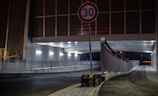 A dimly lit tunnel with an overhead pedestrian walkway, illuminated by industrial lighting. A 30 km/h speed limit sign is prominently displayed in the foreground, and traffic barriers with black and yellow stripes are placed around the signpost.