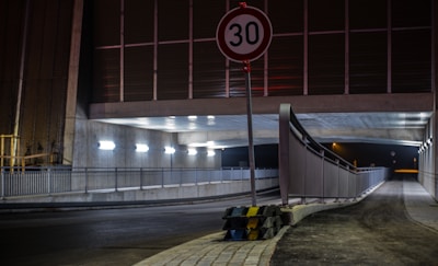 A dimly lit tunnel with an overhead pedestrian walkway, illuminated by industrial lighting. A 30 km/h speed limit sign is prominently displayed in the foreground, and traffic barriers with black and yellow stripes are placed around the signpost.