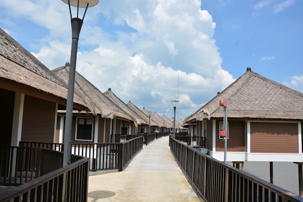 A row of bungalows with thatched roofs lines both sides of a long pathway elevated over water. Each bungalow has wooden railings and is designed with brown and white colors. The sky is partly cloudy, and there are street lamps along the pathway.