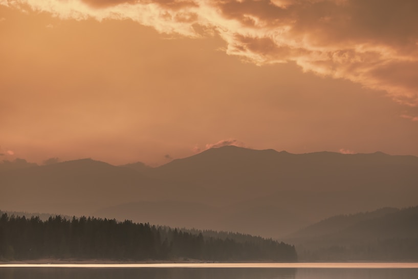A serene landscape featuring a mountain range under a golden sky filled with dramatic clouds. The foreground includes a calm body of water reflecting the light, bordered by a line of trees. The soft hues create a tranquil and majestic atmosphere.