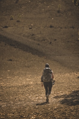 A person with a large backpack walks alone on a barren, rocky landscape under a golden sunlight. The terrain appears dry and sparse, with few visible plants.