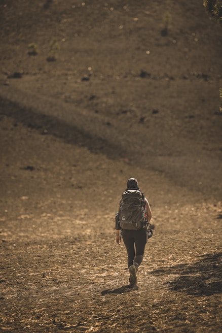 A person with a large backpack walks alone on a barren, rocky landscape under a golden sunlight. The terrain appears dry and sparse, with few visible plants.