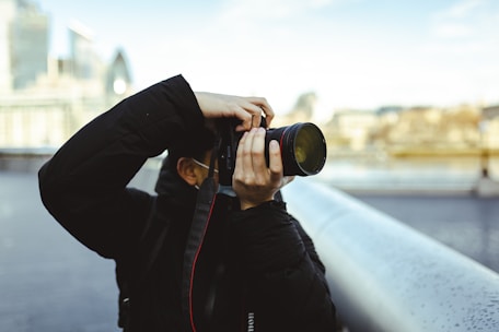 A person dressed in a dark jacket is holding a professional camera, taking a photograph. The background is a blurred cityscape featuring modern buildings and a bridge.