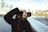 A person dressed in a dark jacket is holding a professional camera, taking a photograph. The background is a blurred cityscape featuring modern buildings and a bridge.