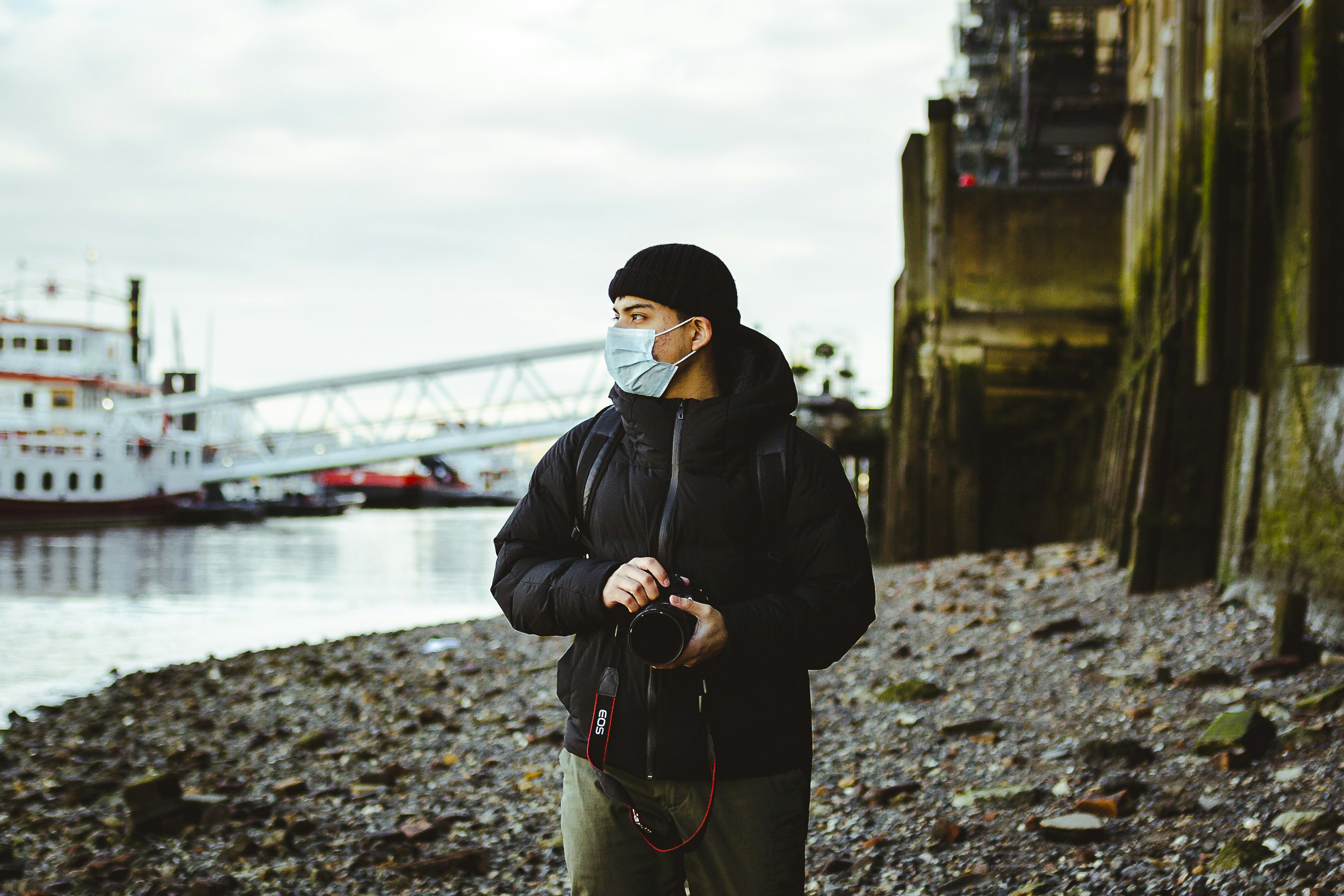 Man in mask holding camera near river with boat