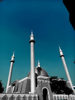 A large mosque with a prominent dome and several tall minarets under a clear blue sky. The architecture features intricate patterns and a blend of traditional and modern elements. Trees partially frame the structure, adding contrast to the urban setting.