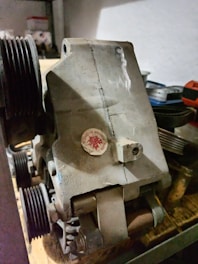 A mechanical component with multiple pulleys is placed on a shelf. The unit appears worn and dusty, suggesting it has been stored or unused for some time. There is a red sticker affixed to its surface, possibly a logo or certification mark.