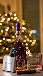 A glass of Cognac on a wooden table beside an open book and a small plate of chocolates.