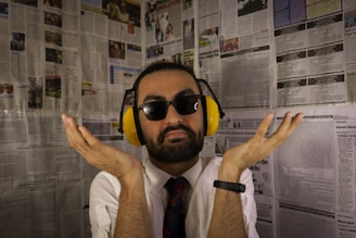 A man with a beard and sunglasses is wearing yellow noise-cancelling headphones. He is dressed in a white shirt and colorful tie, with a black smartwatch on his left wrist. The background consists of walls covered in newspaper pages.
