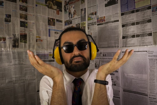 A man with a beard and sunglasses is wearing yellow noise-cancelling headphones. He is dressed in a white shirt and colorful tie, with a black smartwatch on his left wrist. The background consists of walls covered in newspaper pages.