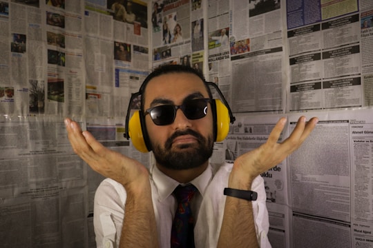 A man with a beard and sunglasses is wearing yellow noise-cancelling headphones. He is dressed in a white shirt and colorful tie, with a black smartwatch on his left wrist. The background consists of walls covered in newspaper pages.