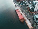 An aerial view depicts a cargo ship docked at a port beside an urban area. The vessel, characterized by its large red containers, is adjacent to industrial buildings and city infrastructure. The surrounding water is a deep shade of blue, contrasting with the organized layout of the streets and buildings.