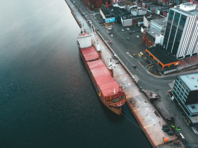 An aerial view depicts a cargo ship docked at a port beside an urban area. The vessel, characterized by its large red containers, is adjacent to industrial buildings and city infrastructure. The surrounding water is a deep shade of blue, contrasting with the organized layout of the streets and buildings.
