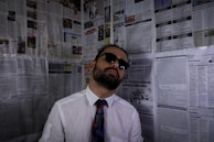 A person wearing sunglasses, a white shirt, and a colorful tie leans against a wall covered with newspaper pages. The individual has a beard and an expression of indifference or disinterest.