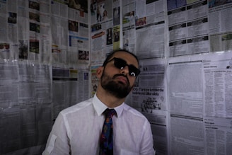 A person wearing sunglasses, a white shirt, and a colorful tie leans against a wall covered with newspaper pages. The individual has a beard and an expression of indifference or disinterest.