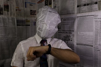 A person with their head wrapped in a newspaper sits in a room covered in newspaper pages. They are wearing a white shirt with a tie and looking at a wristwatch. The atmosphere seems surreal and thought-provoking.