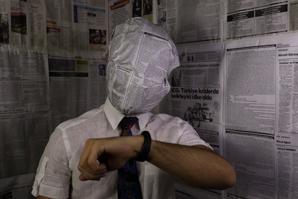 A person with their head wrapped in a newspaper sits in a room covered in newspaper pages. They are wearing a white shirt with a tie and looking at a wristwatch. The atmosphere seems surreal and thought-provoking.