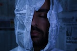 A person with their eyes closed is enveloped in newspaper sheets, creating a surreal or artistic scene. The background also contains newspaper pages, giving a sense of being surrounded by text and print.
