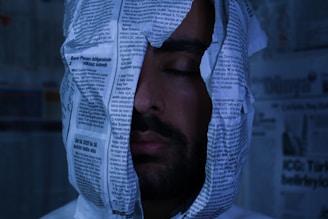 A person with their eyes closed is enveloped in newspaper sheets, creating a surreal or artistic scene. The background also contains newspaper pages, giving a sense of being surrounded by text and print.