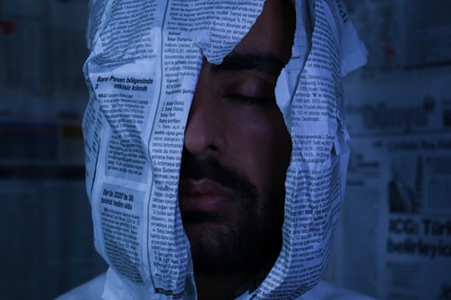 A person with their eyes closed is enveloped in newspaper sheets, creating a surreal or artistic scene. The background also contains newspaper pages, giving a sense of being surrounded by text and print.