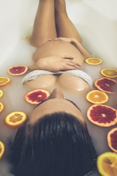 A pregnant woman is partially submerged in a bathtub filled with milky water, surrounded by slices of citrus fruits like oranges and grapefruits. She is wearing white lace clothing and resting her hands on her belly, creating a serene and intimate atmosphere.