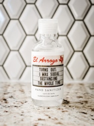 A bottle of hand sanitizer placed on a speckled countertop in front of a hexagonal tile backsplash. The label on the bottle features the text 'El Arroyo' in red and a humorous message about social distancing.