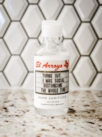 A bottle of hand sanitizer placed on a speckled countertop in front of a hexagonal tile backsplash. The label on the bottle features the text 'El Arroyo' in red and a humorous message about social distancing.