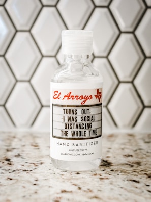 A bottle of hand sanitizer placed on a speckled countertop in front of a hexagonal tile backsplash. The label on the bottle features the text 'El Arroyo' in red and a humorous message about social distancing.
