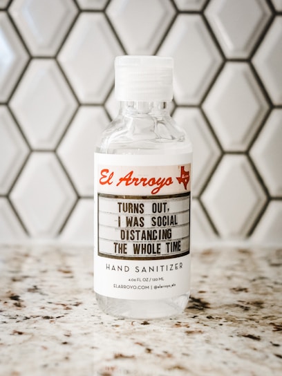 A bottle of hand sanitizer placed on a speckled countertop in front of a hexagonal tile backsplash. The label on the bottle features the text 'El Arroyo' in red and a humorous message about social distancing.