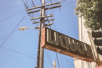A vintage liquor store sign hangs prominently with bold lettering. It is attached to a weathered building, with a utility pole and power lines visible in the background. The sky is clear and blue, and a traffic light is partially in view at the right side of the image.