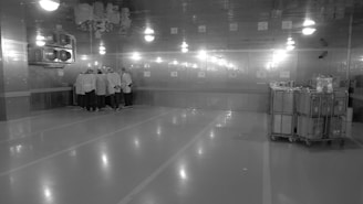 A large, industrial-looking room with reflective walls and ceiling. A group of people wearing protective clothing and hair coverings are gathered in a cluster near the corner. The room is well-lit, with bright overhead lights, and it appears to be a controlled or sterile environment. On the right side, there are metal carts which seem to hold equipment or supplies.