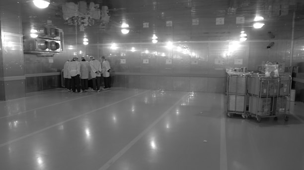 A large, industrial-looking room with reflective walls and ceiling. A group of people wearing protective clothing and hair coverings are gathered in a cluster near the corner. The room is well-lit, with bright overhead lights, and it appears to be a controlled or sterile environment. On the right side, there are metal carts which seem to hold equipment or supplies.