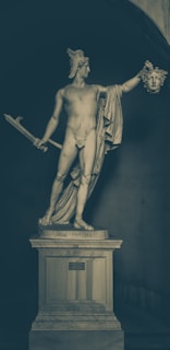 A classical statue of a nude male figure, adorned with a winged helmet, holding a sword in one hand and the head of Medusa in the other. It stands on a pedestal within an archway, capturing the dramatic pose from Greek mythology.