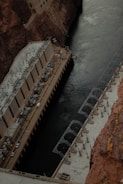 A large concrete structure with intricate machinery is built into a canyon, adjacent to a dark river. The structure appears to be a power plant or industrial facility, with various pipes and electrical components visible on its roof. The rocky walls of the canyon tower above, providing a stark contrast to the smooth surface of the water below.