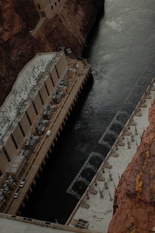 A large concrete structure with intricate machinery is built into a canyon, adjacent to a dark river. The structure appears to be a power plant or industrial facility, with various pipes and electrical components visible on its roof. The rocky walls of the canyon tower above, providing a stark contrast to the smooth surface of the water below.