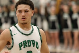 A basketball player is wearing a white jersey with green accents and the word 'Provo' on the front. The player has short curly hair and is in focus, while the background is blurred with indistinct figures that appear to be cheerleaders in black and white uniforms.