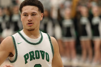 A basketball player is wearing a white jersey with green accents and the word 'Provo' on the front. The player has short curly hair and is in focus, while the background is blurred with indistinct figures that appear to be cheerleaders in black and white uniforms.