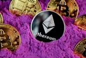 A collection of cryptocurrency coins, including Ethereum and Bitcoin, are partially buried in bright purple sand. The Ethereum coin is prominently featured at the center, while Bitcoin coins surround it.
