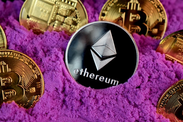 A collection of cryptocurrency coins, including Ethereum and Bitcoin, are partially buried in bright purple sand. The Ethereum coin is prominently featured at the center, while Bitcoin coins surround it.
