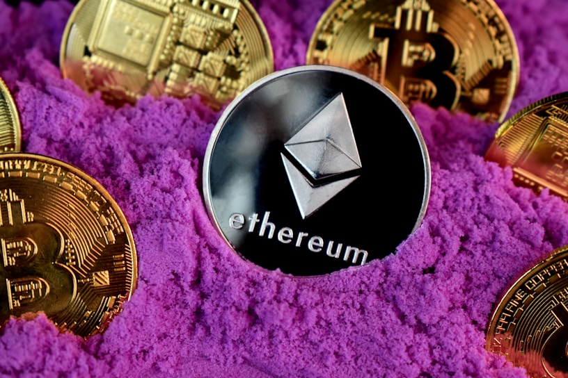A collection of cryptocurrency coins, including Ethereum and Bitcoin, are partially buried in bright purple sand. The Ethereum coin is prominently featured at the center, while Bitcoin coins surround it.