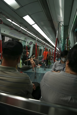 A subway train interior with people seated and standing, holding onto red handrails. Overhead fluorescent lights illuminate the space, and some passengers are wearing masks.