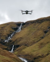 A drone hovers in the air against a backdrop of rolling hills with a small waterfall cascading down the rocky terrain.
