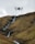 A drone hovers in the air against a backdrop of rolling hills with a small waterfall cascading down the rocky terrain.