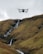 A drone hovers in the air against a backdrop of rolling hills with a small waterfall cascading down the rocky terrain.