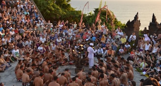 A large group of people seated in a circular formation around performers, with lush greenery and ocean visible in the background. The performers are wearing traditional attire and the audience appears engaged, with some taking photos. Decorative elements and traditional architecture are visible.