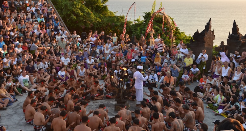 A large group of people seated in a circular formation around performers, with lush greenery and ocean visible in the background. The performers are wearing traditional attire and the audience appears engaged, with some taking photos. Decorative elements and traditional architecture are visible.